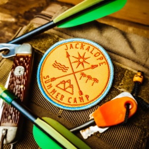 Op de afbeelding: Een geborduurde patch in geel en oranje met de tekst "Jackalope Summer Camp" en een kompasontwerp met een zon, golven, bergen en palmbomen. De patch bevindt zich op een bruine canvas tas met een groene pijl, een multitoolmes en een klein oranje gereedschap.