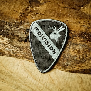May include: A black and silver embroidered patch with a white deer head and the text "1st DIVISION".