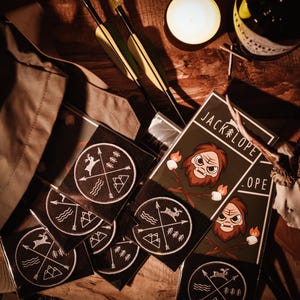 May include: A collection of black and white embroidered patches featuring a compass design and a Jackalope logo. The patches are displayed on a wooden surface alongside a brown leather bag, arrows, and a candle.