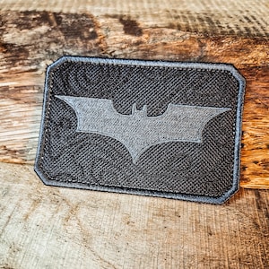 Puede incluir: Un parche negro bordado con una silueta de murciélago gris. El parche es rectangular y tiene un borde cosido.