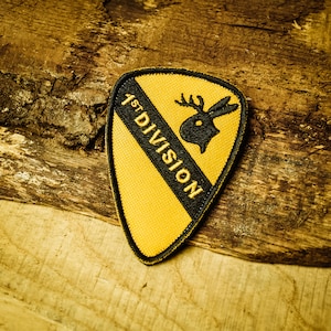 Op de afbeelding: Een gele en zwarte patch met de tekst "1st DIVISION" en een gestileerd hertenhoofd met geweien.