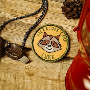 Könnte beinhalten: Ein gestickter Aufnäher in Gelb und Grün mit einem Cartoon-Waschbären und dem Text "Jackalope Camp Luke".