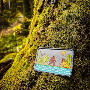 May include: A rectangular patch featuring a Bigfoot design in a forest scene. The patch includes a mountain range, trees, a tent, and a sun. The Bigfoot is brown, the trees are green, and the water is turquoise. The patch is set against a gray background.