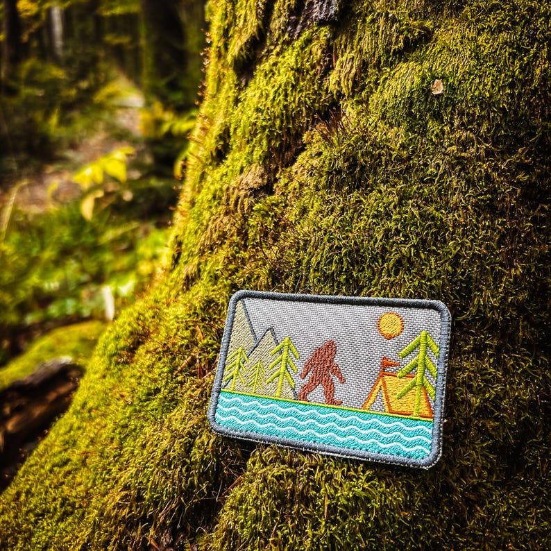 May include: A rectangular patch featuring a Bigfoot design in a forest scene. The patch includes a mountain range, trees, a tent, and a sun. The Bigfoot is brown, the trees are green, and the water is turquoise. The patch is set against a gray background.