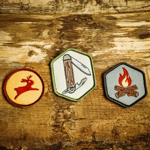 Op de afbeelding: Drie geborduurde patches op een houten achtergrond. De patches zijn zeshoekig van vorm en tonen een rood hert, een zakmes en een kampvuur met vlammen.