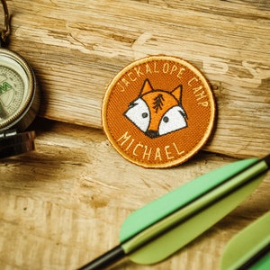 Op de afbeelding: Een bruin geborduurd patch met een vos-ontwerp en de tekst "Jackalope Camp Michael".