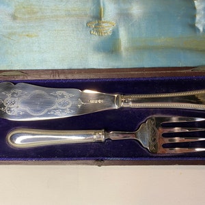 Puede incluir: Un juego de cuchillo y tenedor de mantequilla de plata en una caja forrada de terciopelo azul. El cuchillo y el tenedor tienen diseños ornamentados y están en excelente estado.