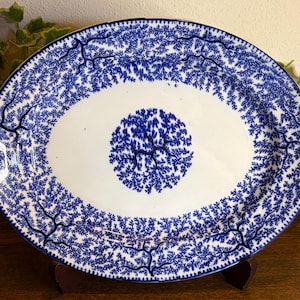 May include: Oval serving platter with a white centre and a blue tree branch pattern. The rim and centre of the platter feature a detailed blue design. The platter is displayed on a wooden stand.