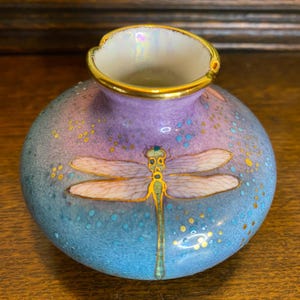 Puede incluir: Un jarrón pequeño y redondo con un degradado de tonos azules y morados. Un diseño detallado de libélula está centrado en el jarrón. El borde está adornado con oro. El jarrón es probablemente de cerámica o porcelana.