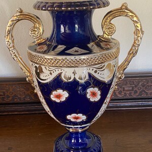 Pode incluir: Um vaso de porcelana azul e branco com detalhes dourados e desenhos florais. O vaso tem duas al&ccedil;as douradas ornamentadas e uma base quadrada.