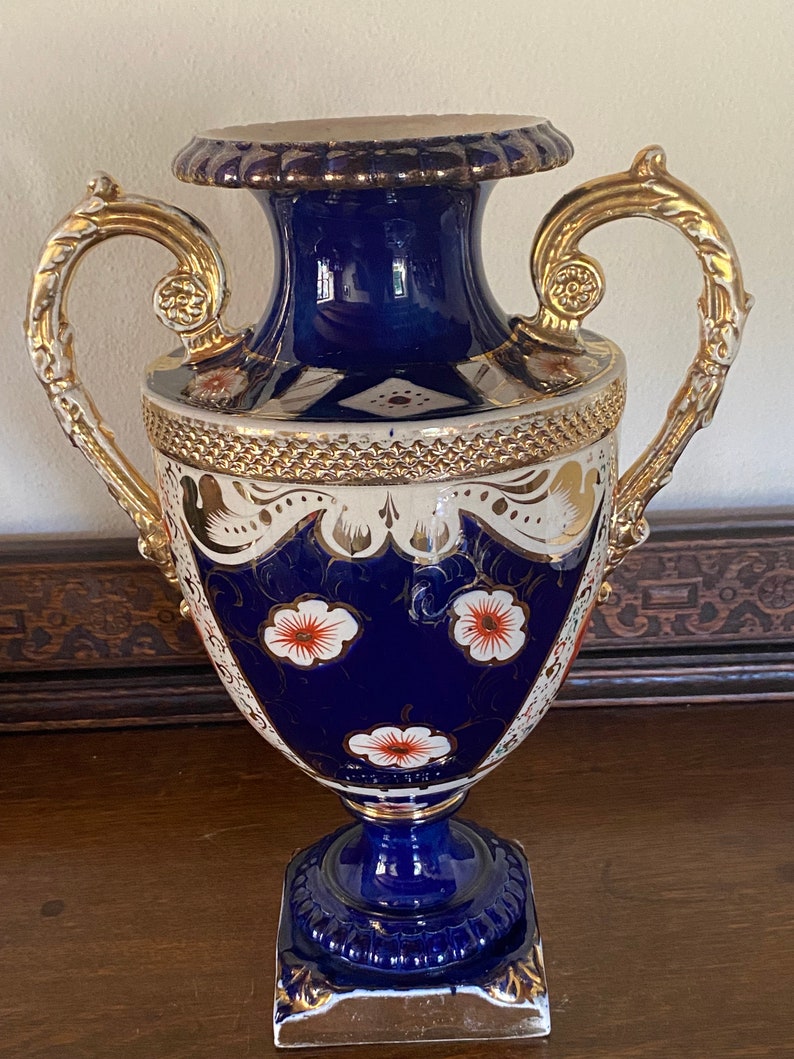 Pode incluir: Um vaso de porcelana azul e branco com detalhes dourados e desenhos florais. O vaso tem duas al&ccedil;as douradas ornamentadas e uma base quadrada.