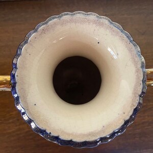 Pode incluir: Um vaso de cer&acirc;mica branca com al&ccedil;as douradas e uma borda azul e dourada. O vaso tem uma borda festonada e uma abertura larga.