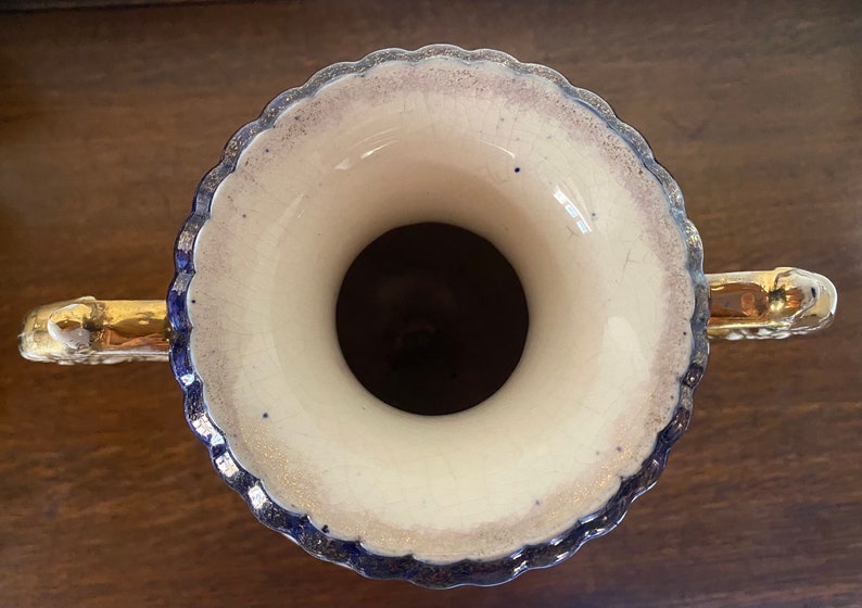 Pode incluir: Um vaso de cer&acirc;mica branca com al&ccedil;as douradas e uma borda azul e dourada. O vaso tem uma borda festonada e uma abertura larga.