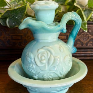 May include: A light blue, vintage-style candle holder with a rose design. The candle holder has a small handle and sits in a matching dish. The color transitions from light blue to a darker teal.