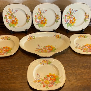 May include: Set of seven vintage floral patterned plates, including a large oval serving platter. The plates are cream-coloured with a gold trim and feature a floral design with orange, yellow, and pink flowers.