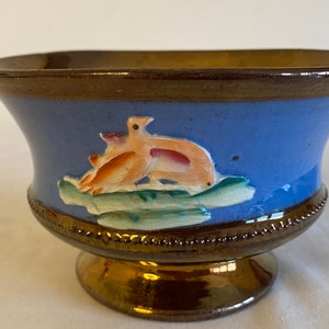 Puede incluir: Un pequeño cuenco con pie, con un esmalte azul y un diseño pintado de dos pájaros sobre una hoja verde. El cuenco tiene un borde y una base dorados.