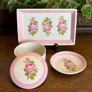 Könnte beinhalten: Ein Set aus Vintage-Keramik-Servierteilen mit einem floralen Rosendesign. Das Set umfasst ein rechteckiges Tablett, einen runden Behälter mit Deckel und eine kleine Schale. Die Teile sind weiß mit rosa Akzenten und Rosenillustrationen.