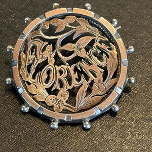 May include: A circular brooch featuring a silver outer rim with small spherical accents and a central rose gold-toned cutout design. The intricate metalwork displays floral motifs and the name FLORENCE in stylized lettering across the center.
