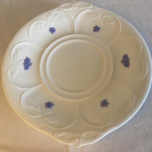May include: A white porcelain saucer with a floral design. The saucer has a scalloped edge and a center well. The floral design is in shades of blue and features small clusters of flowers.