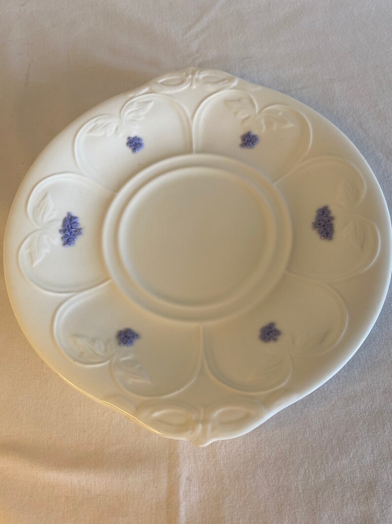 May include: A white porcelain saucer with a floral design. The saucer has a scalloped edge and a center well. The floral design is in shades of blue and features small clusters of flowers.