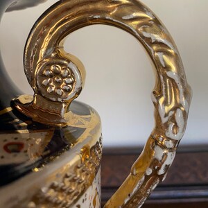 Pode incluir: Um vaso de cer&acirc;mica branco e dourado com um design floral detalhado e uma al&ccedil;a dourada. O vaso tem uma base azul e branca com detalhes dourados.