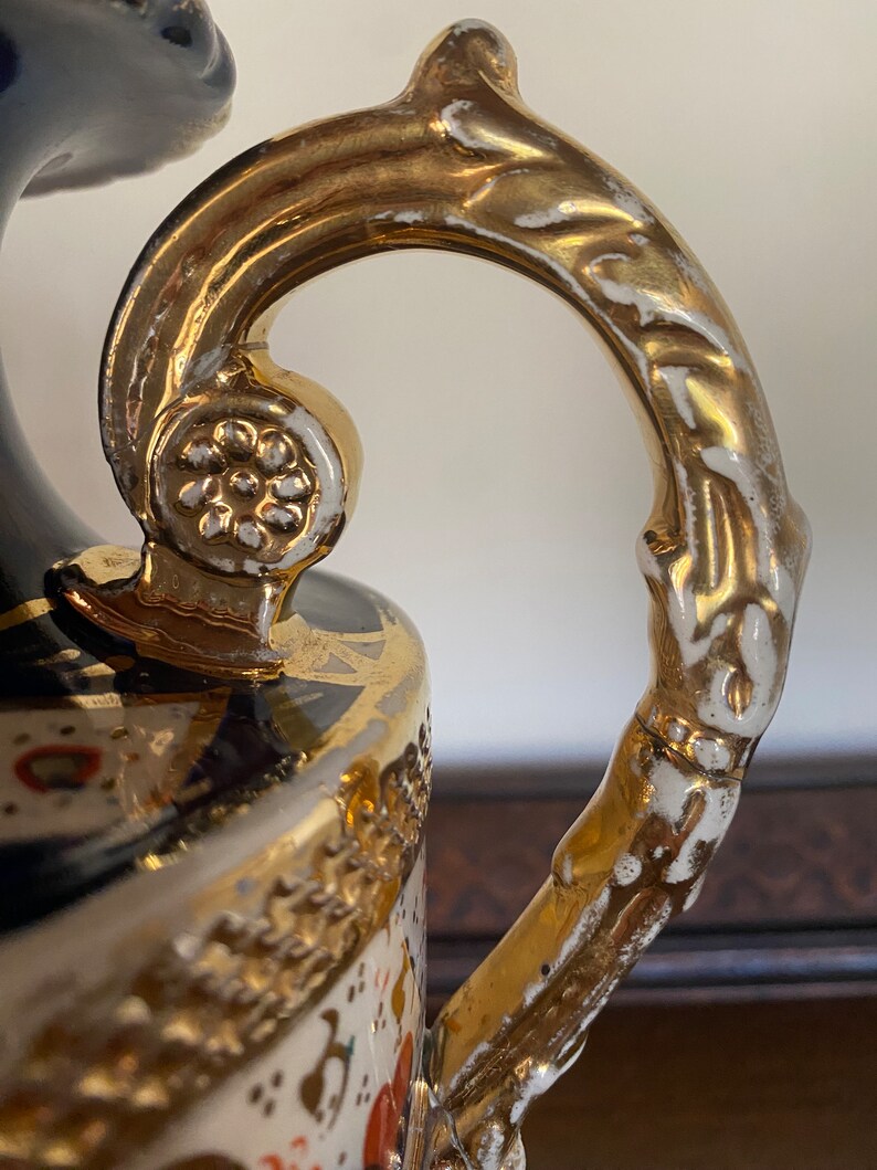 Pode incluir: Um vaso de cer&acirc;mica branco e dourado com um design floral detalhado e uma al&ccedil;a dourada. O vaso tem uma base azul e branca com detalhes dourados.