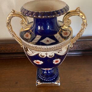 Pode incluir: Um vaso de porcelana azul e branco com detalhes dourados e desenhos florais. O vaso tem duas al&ccedil;as douradas ornamentadas e uma base quadrada.
