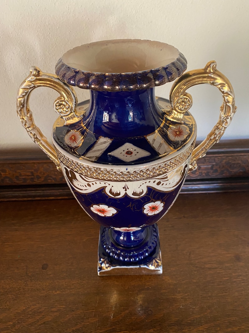 Pode incluir: Um vaso de porcelana azul e branco com detalhes dourados e desenhos florais. O vaso tem duas al&ccedil;as douradas ornamentadas e uma base quadrada.