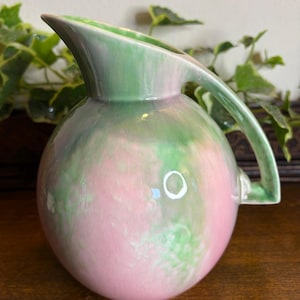 May include: A vintage ceramic pitcher with a round body and a curved handle. The pitcher is painted with a blend of pink and green hues, creating a soft, watercolour effect. The spout is pointed and the handle is a matching green.