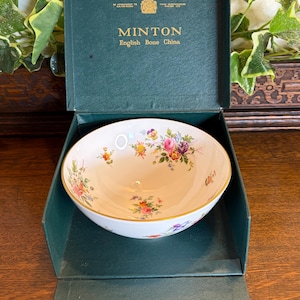 May include: A white Minton English bone china bowl featuring a delicate floral pattern with pink, yellow, and purple flowers and a gold rim. The bowl is presented inside a dark green box with gold text that reads MINTON English Bone China.