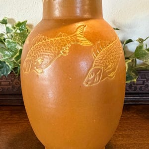 May include: A tall, terracotta-coloured ceramic vase featuring a smooth, rounded body and a flared rim. The exterior surface is decorated with two embossed fish motifs and a repeating scalloped pattern with small dots encircling the neck area.