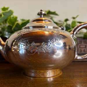 May include: A polished silver-tone teapot featuring a rounded body, a curved handle, a short spout, and a lid with a small finial. The upper portion of the teapot is decorated with an engraved scrollwork pattern that encircles the main body.