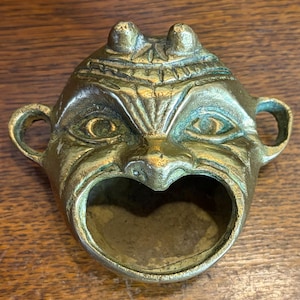 May include: A brass bottle opener in the shape of a stylized face with an open mouth. The opener has a detailed design with eye sockets, a nose, and two small handles on either side. The brass has a patina.