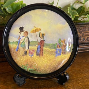 May include: A circular decorative plate with a black frame and stand. The plate displays a pastoral scene with figures in period attire, strolling through a field under a yellow parasol. The artwork has a vintage style.