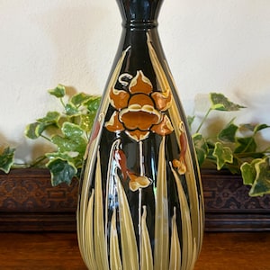 May include: A tall, decorative vase with a flared top and a wide base. The vase is primarily black with a floral design in shades of brown and tan. The design includes stylized flowers and long, slender leaves.