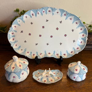 Könnte beinhalten: Ein weißes Keramik-Serviertablett und ein passendes Geschirrset mit blauem und rotem Blumendesign. Das Set umfasst eine große ovale Platte, zwei Deckelgläser und eine kleine Schale. Die Teile sind mit kleinen roten Blumen und grauen Schmetterlingsakzenten verziert.