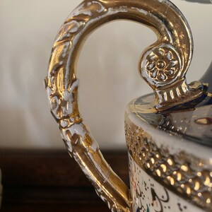 Pode incluir: Um vaso de porcelana branca com uma al&ccedil;a dourada e detalhes dourados intrincados. O vaso tem um padr&atilde;o floral azul e branco.