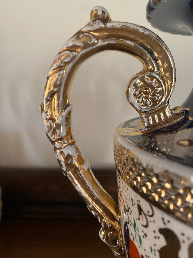 Pode incluir: Um vaso de porcelana branca com uma al&ccedil;a dourada e detalhes dourados intrincados. O vaso tem um padr&atilde;o floral azul e branco.