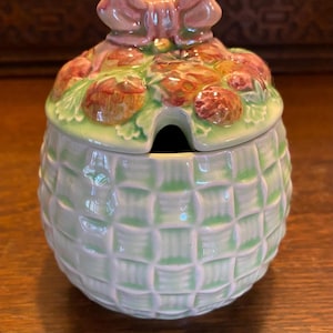 May include: A ceramic sugar bowl with a lid, featuring a light green basketweave design. The lid is adorned with a cluster of red berries and green leaves, topped with a pink bow. A small opening is present for a spoon.