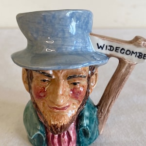 May include: A ceramic figurine of a man with a blue hat and a brown beard. He is wearing a green jacket and a pink shirt. The figurine has a wooden handle with a sign that reads "Widecombe".