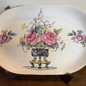 May include: Oval ceramic serving platter with a floral design. The platter features pink roses, yellow flowers, and green leaves. The flowers are arranged in a vase on a decorative stand. The platter has a cream-colored background.