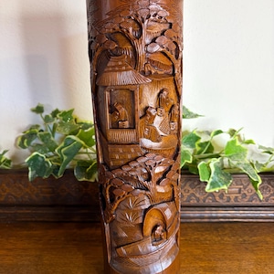 May include: A tall, cylindrical wooden vase with intricate carvings depicting a detailed landscape scene. The dark brown vase features figures, trees, and buildings, showcasing a traditional Asian-inspired design. The vase is displayed on a wooden surface.