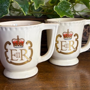 Puede incluir: Dos tazas color crema con borde y asa dorados. Cada taza presenta una corona y las letras "ER" en dorado, con el texto "1952-1977" debajo. Las tazas son probablemente artículos conmemorativos.