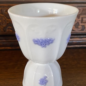 May include: White ceramic egg cup with a lavender floral design. The cup has a fluted base and a wide, rounded top.