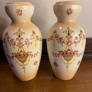 May include: Pair of antique ceramic vases with a cream background and floral designs. The vases have a bulbous body and a narrow neck. The floral designs are in shades of pink, yellow, and green.