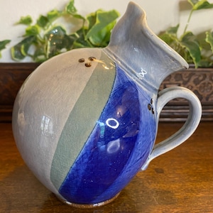 以下が含まれることがあります： ユニークな非対称デザインのセラミックピッチャー。ピッチャーは、注ぎ口とハンドルが付いた丸みを帯びたボディが特徴です。配色は、グレー、ブルー、グリーンのストライプと、ブラウンのアクセントで構成されています。ピッチャーはおそらく飲み物の提供に使用されます。