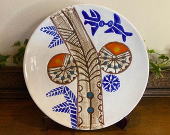 May include: White ceramic decorative plate with a hand-painted design. The plate features a central brown tree-like motif, orange and blue circular patterns, and blue leaf and bird accents. The plate is displayed on a wooden stand.