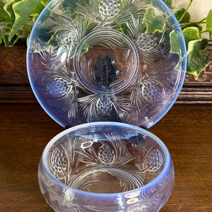 May include: A set of two opalescent blue glass pieces consisting of a plate and a bowl. Both items feature a molded relief pattern of pinecones and pine needles. The glass has a translucent quality with a soft blue tint and a smooth finish.