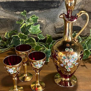 May include: A decorative wine decanter and three glasses. The set has a gold finish with burgundy interiors and floral detailing in white, blue, and green. The decanter has a handle and stopper. The glasses have a stem and a round base, perfect for serving wine.