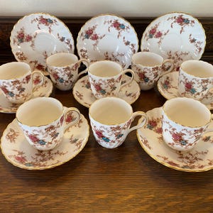 May include: A set of ten teacups and saucers featuring a floral design in shades of pink, blue, and green. The cups and saucers are white with gold edging.